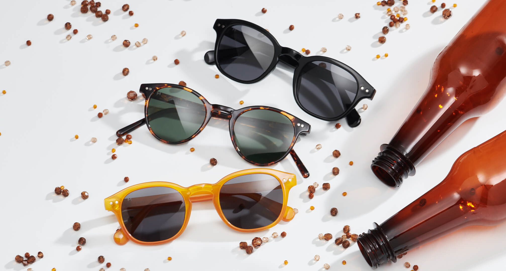 trys saulės akinių poros, pagamintos iš tvarių medžiagų, ant balto stalo, aplinkui karoliukai ir plastikiniai buteliai.
